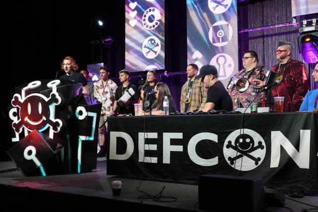 A group of people standing and sitting behind a table with a DEFCON banner, featuring a skull and crossbones logo. The background includes screens displaying similar logos and a person speaking at a podium with a stylized design