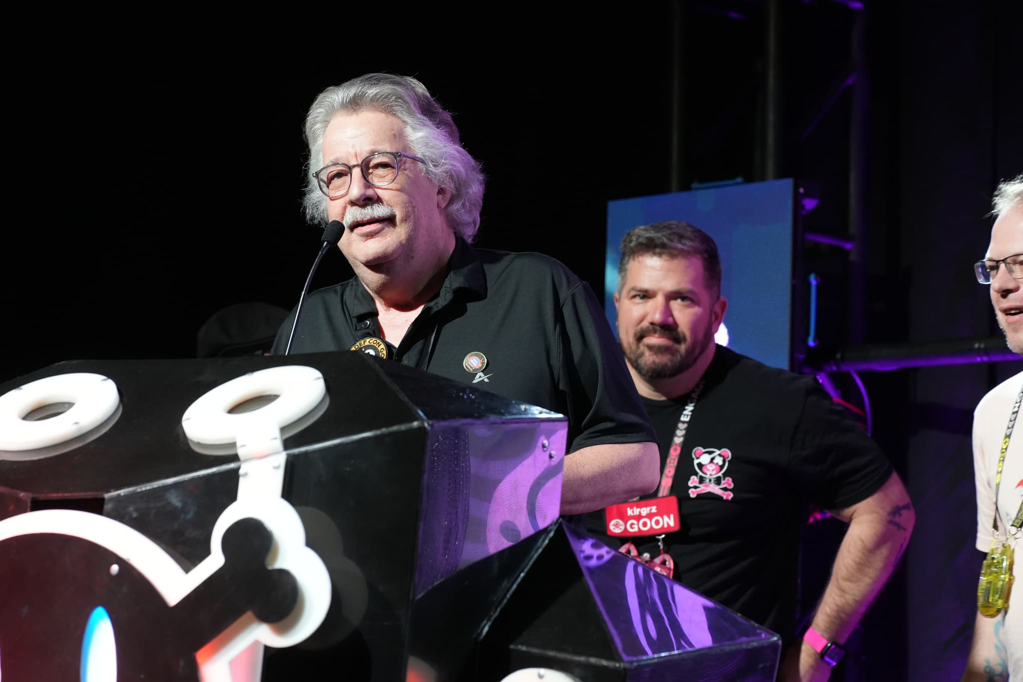 Three men standing on a stage, with one speaking into a microphone at a podium decorated with a stylized skull and crossbones