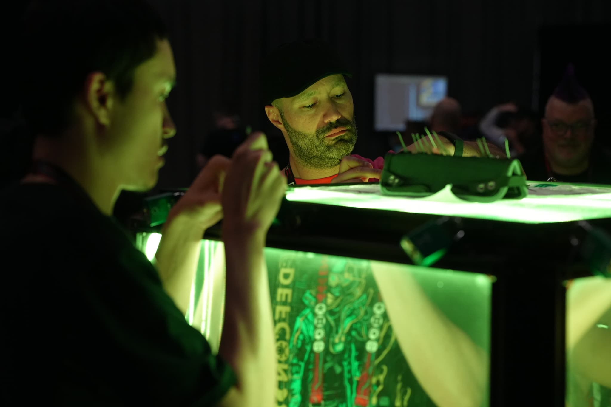 Two people are intently working on a project under green lighting, with one person in the foreground and the other in the background