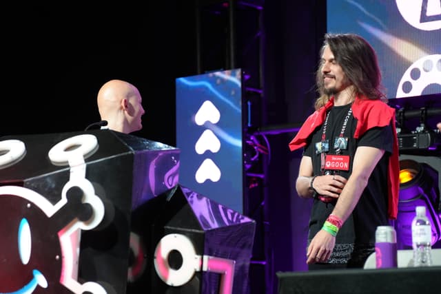 Two individuals on a stage, one standing behind a large, decorated podium and the other wearing a red cape and a black t-shirt with a name tag, with colorful lighting and stage equipment in the background