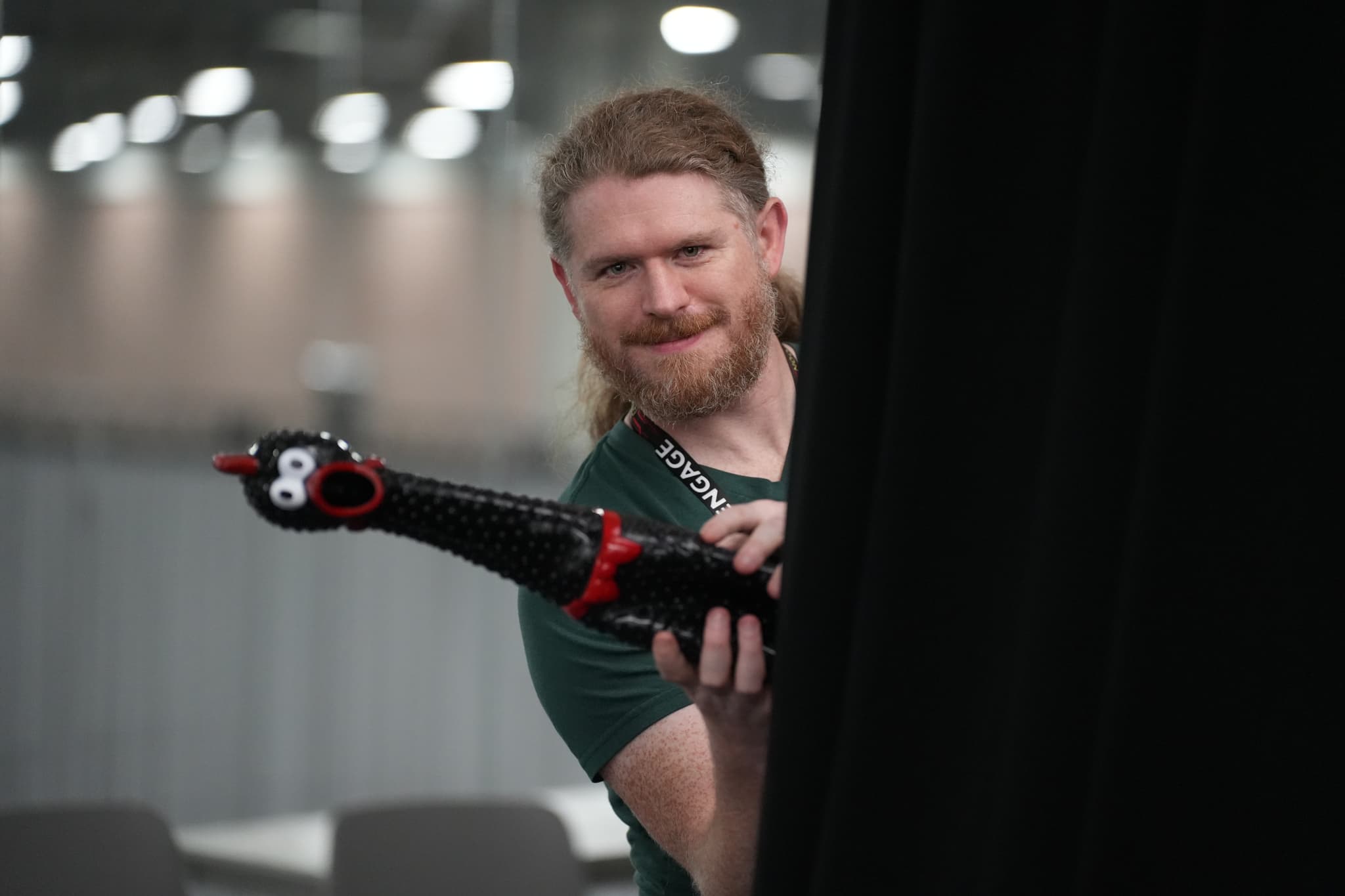 A person with long hair and a beard holding a black rubber chicken with red accents, peeking from behind a black curtain in a well-lit indoor setting