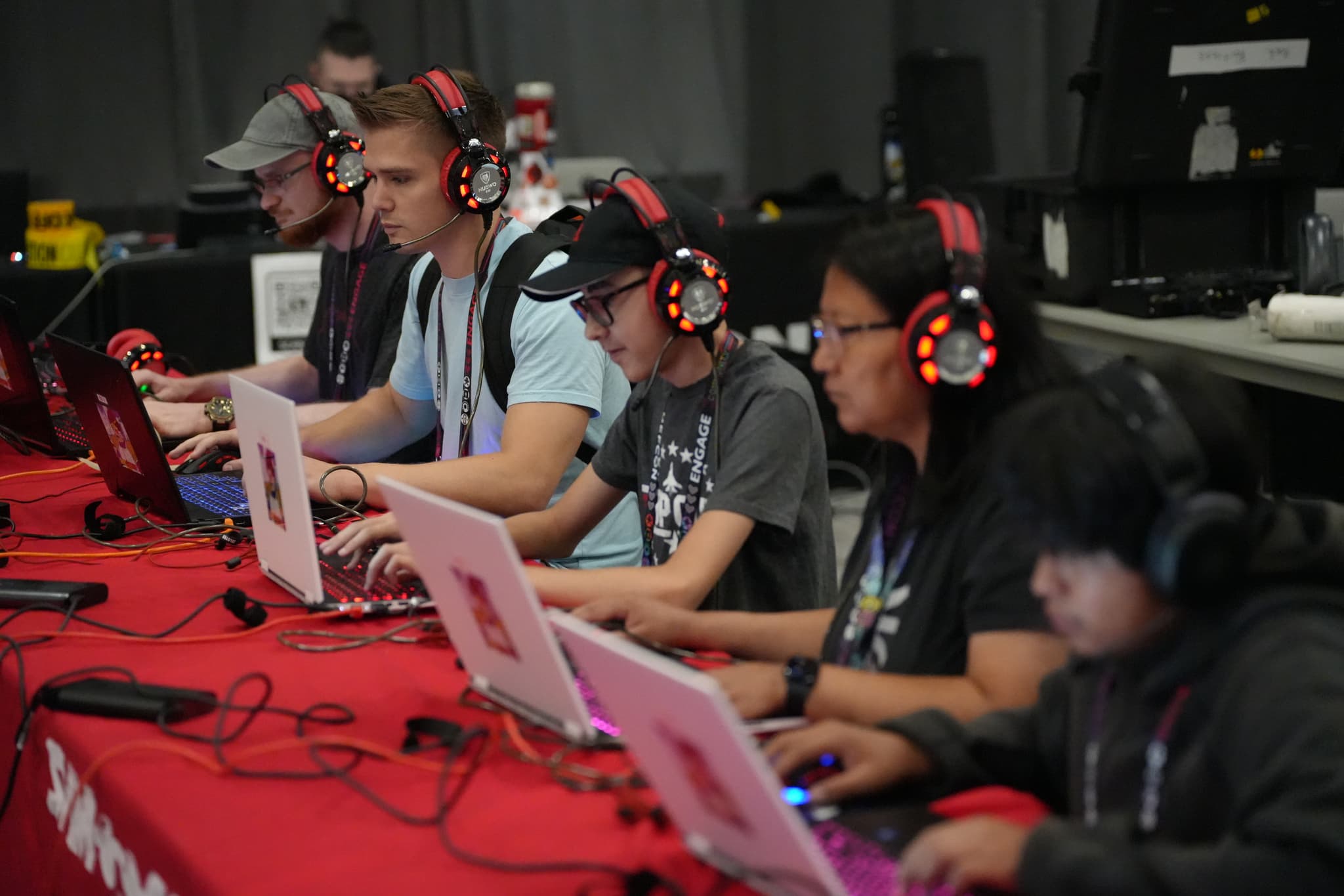 A group of people wearing headphones and using laptops, seated at a table covered with a red tablecloth, likely participating in a gaming or coding event