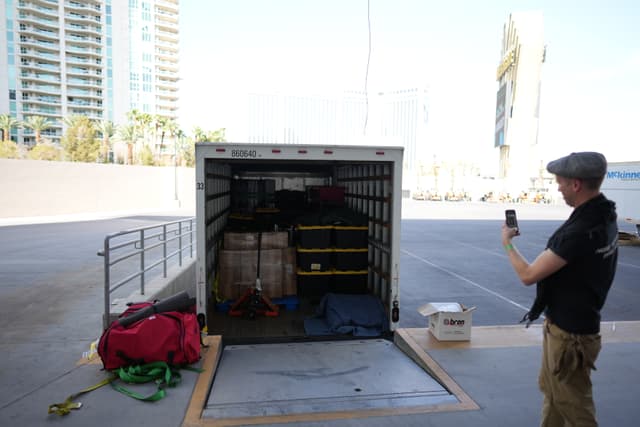 An open truck trailer with various items inside, a person standing nearby taking a photo with a smartphone, and buildings in the background