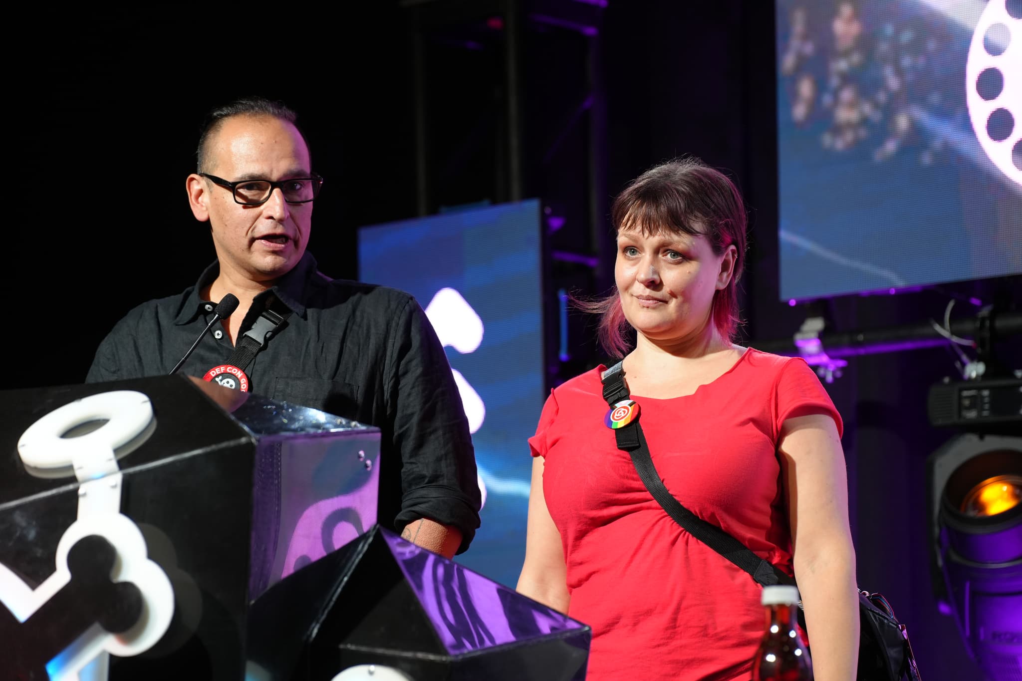 Two people standing on a stage, one speaking into a microphone and the other listening, with a backdrop featuring abstract designs and lighting