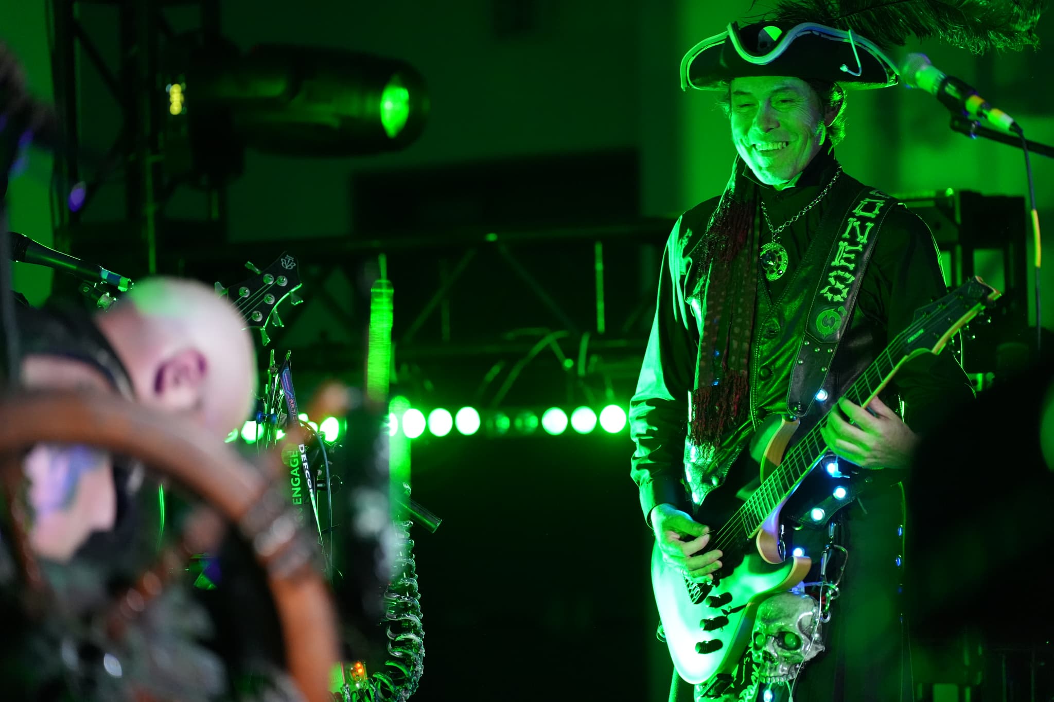 Two musicians performing on stage, one playing a guitar and wearing a pirate hat, illuminated by green lighting