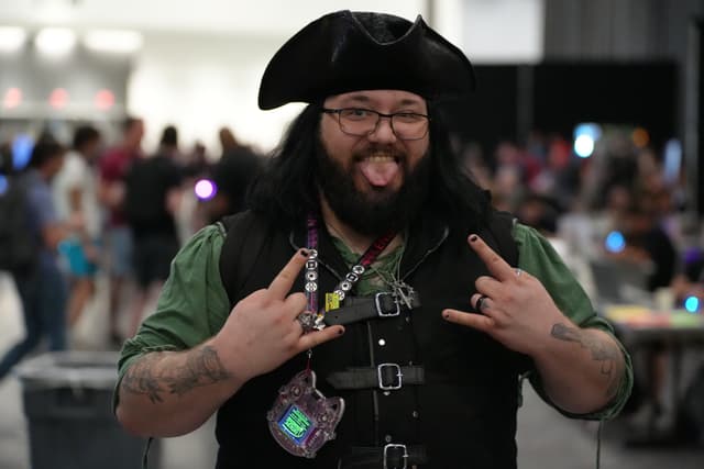 A person dressed in a pirate costume, making a playful gesture with their tongue out and hands forming rock and roll signs, standing in a crowded indoor setting