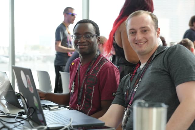 Two people sitting at a table with laptops, smiling at the camera, in a bright room with large windows. Other individuals are visible in the background