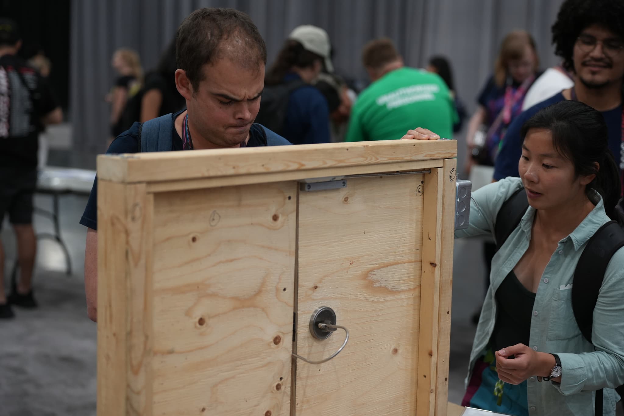 Two people examining a wooden box at an indoor event, with other attendees in the background