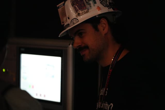 A person wearing a hard hat and a lanyard, standing in a dimly lit environment near a screen displaying bright content