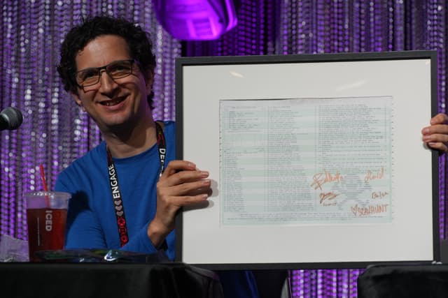 A person with curly hair and glasses, wearing a blue shirt, is smiling and holding a framed document with signatures. The background features a sparkly curtain and purple lighting