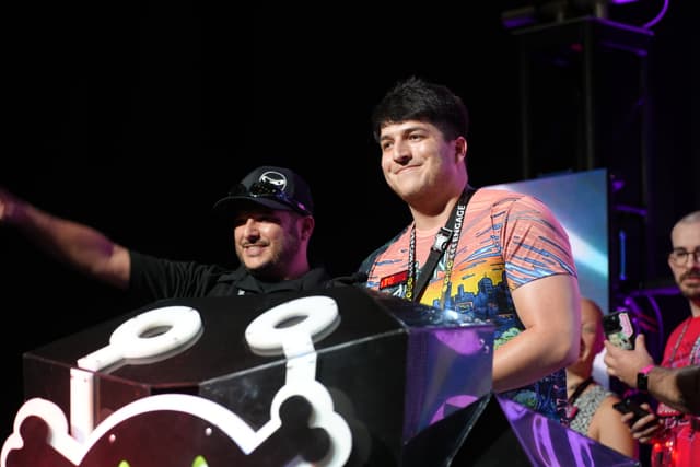 Two people standing on a stage, one wearing a black shirt and cap, the other in a colorful shirt with a lanyard, both smiling and standing behind a podium with a cartoonish design