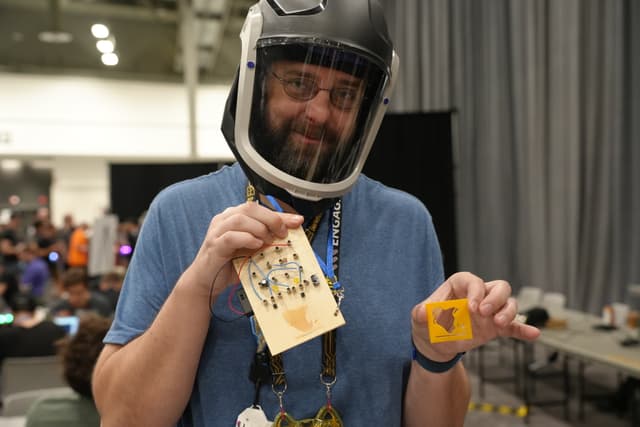 A person wearing a helmet and a blue shirt holds up a small object in each hand, one of which appears to be a card with various items attached to it. The background includes a room with tables and people