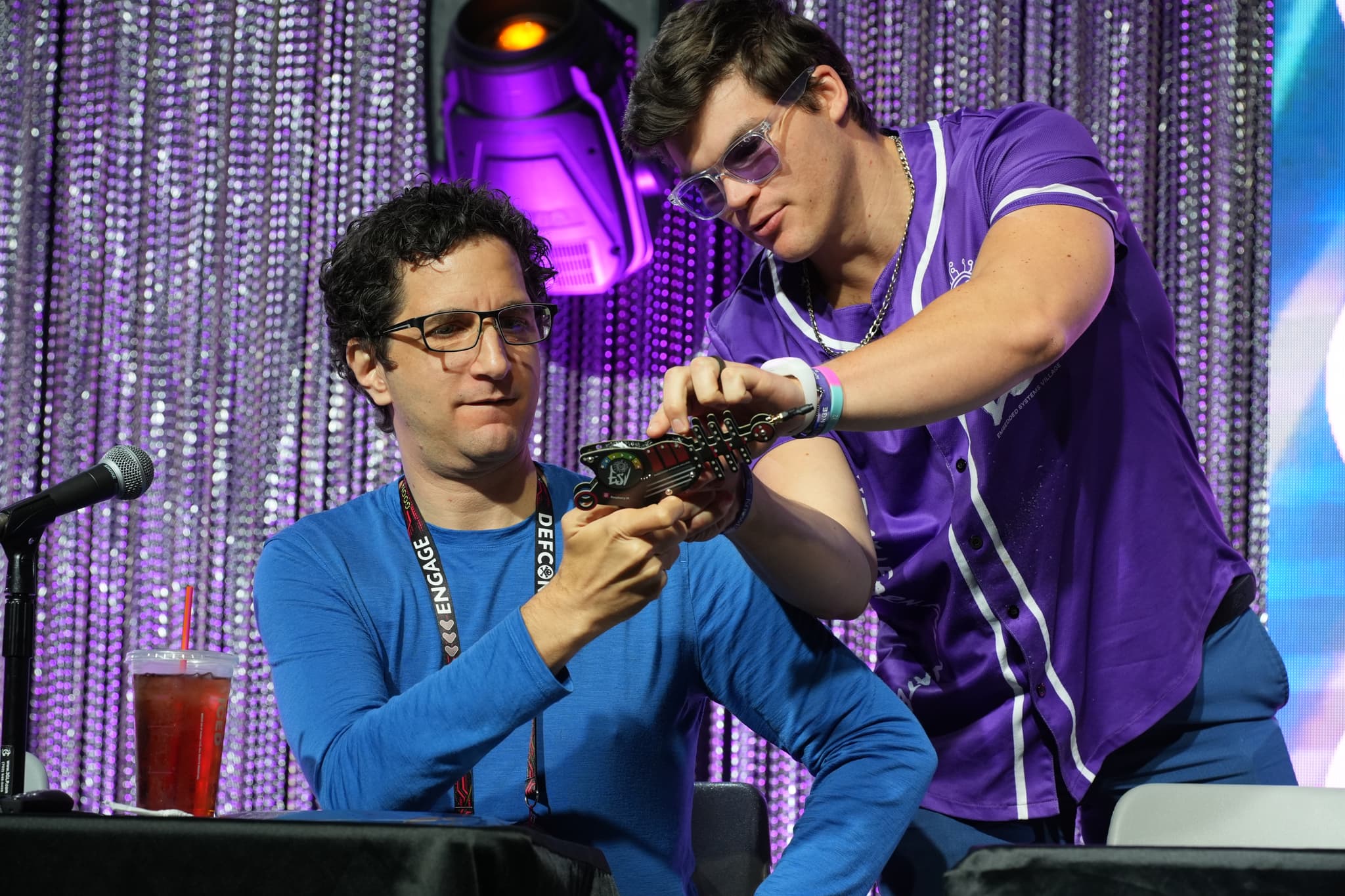 Two individuals are interacting at a table with a microphone and a drink. One person in a blue shirt is seated, while the other in a purple shirt is standing and handing an object to the seated person. The background features a sparkly curtain and colorful lighting