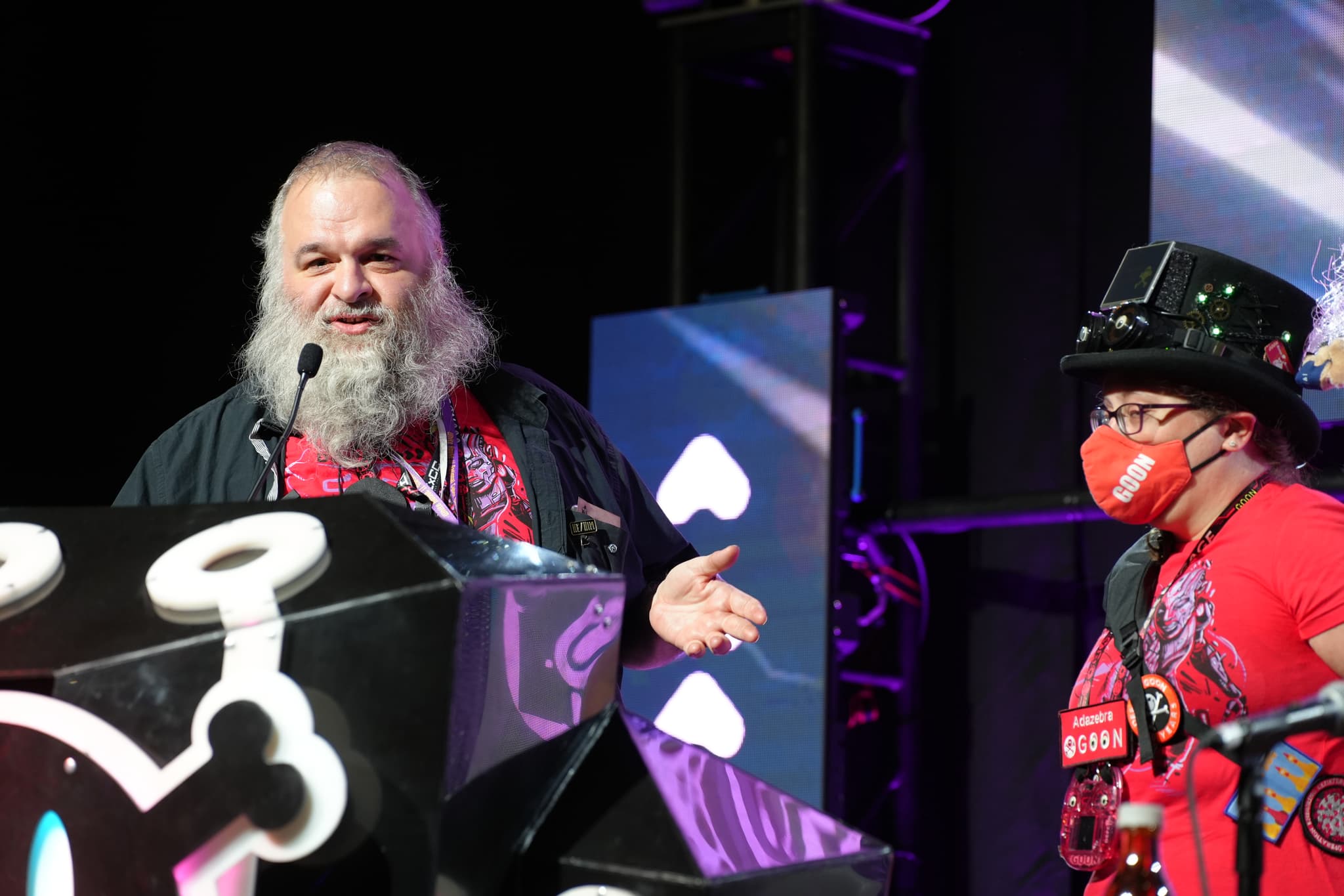 Two individuals on a stage, one speaking at a podium with a microphone and the other wearing a mask and hat, both dressed in red shirts