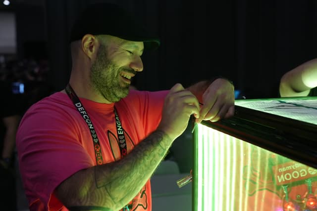 A man wearing a pink shirt and a black cap is smiling and interacting with a brightly lit display case