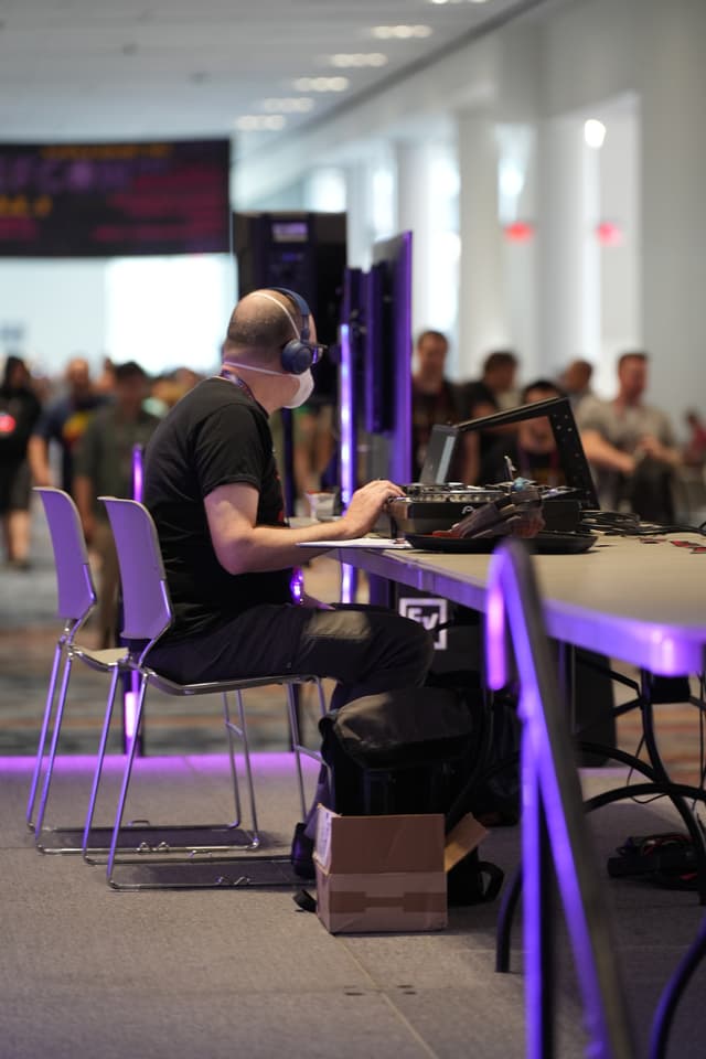 A person wearing headphones is seated at a table with a computer monitor, surrounded by other people in a large, well-lit room