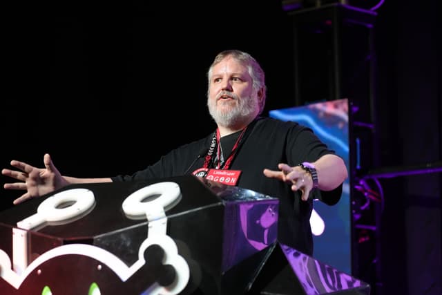 A person standing behind a podium with a colorful design, gesturing with both hands, and wearing a lanyard around their neck