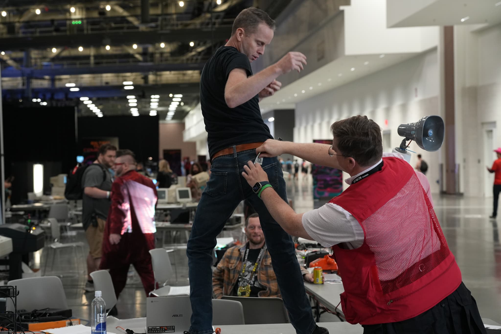 A person in a red vest is playfully grabbing the waistband of another person's pants in a lively indoor setting with several people and tables in the background