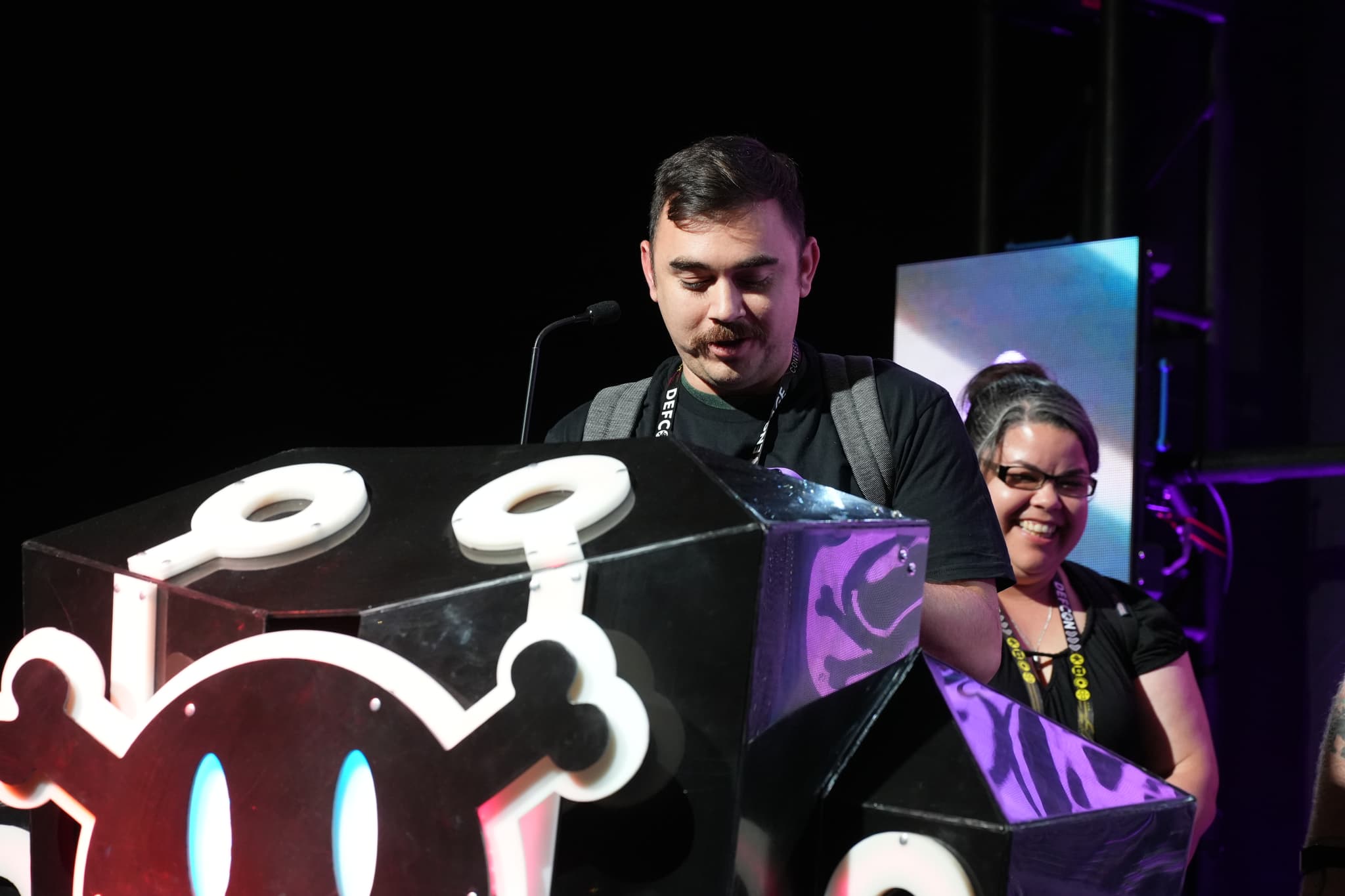 A man standing behind a podium with a stylized skull design, speaking into a microphone, while a woman stands nearby smiling