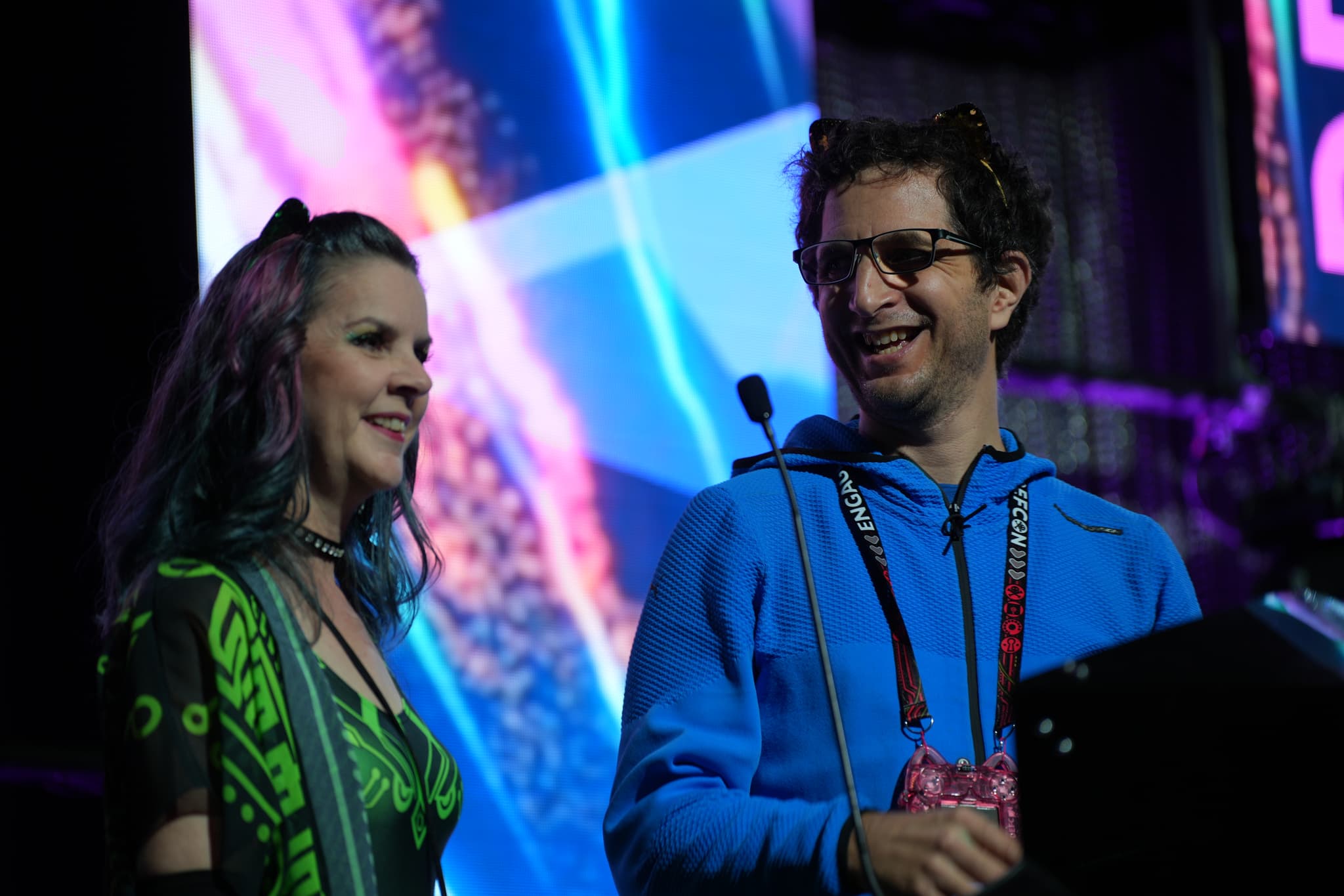 Two people standing together, smiling and talking, with a colorful, abstract background behind them. One person is wearing a green patterned outfit, and the other is in a blue hoodie with glasses