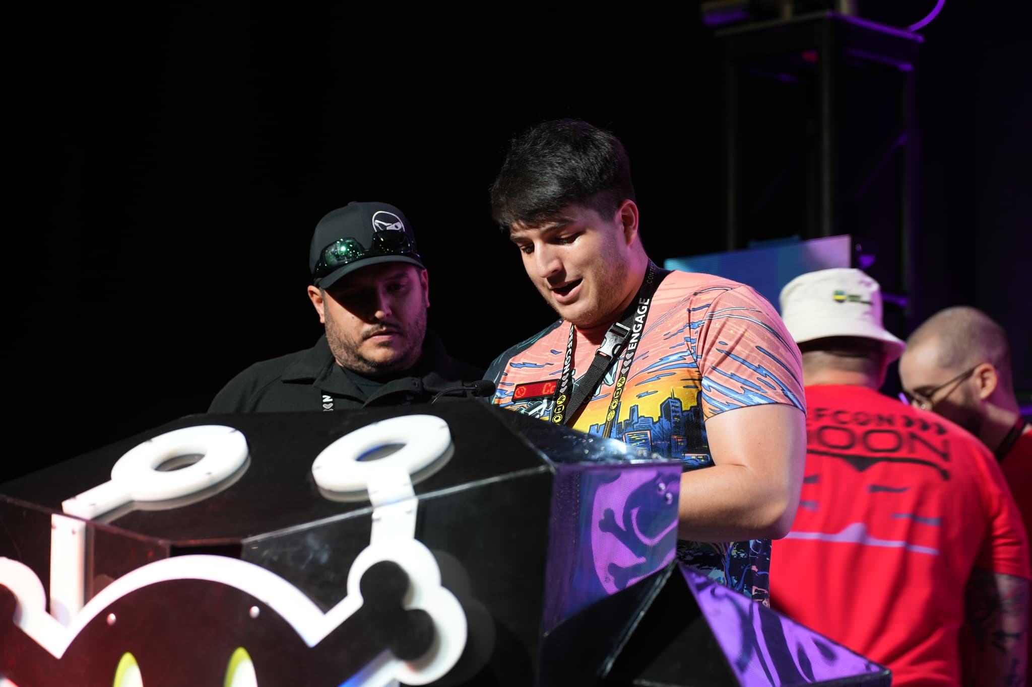 Two men are standing behind a podium with a cartoonish design, engaged in conversation. One man is wearing a colorful shirt, while the other is dressed in black. Two additional individuals are visible in the background, one in a red shirt and the other in a white hat