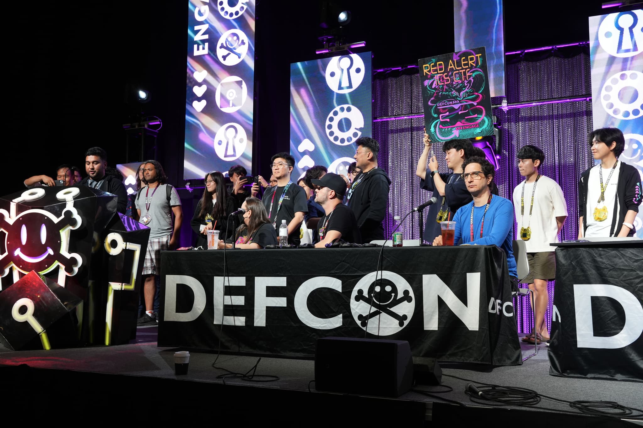 A group of people standing and sitting behind a table with a DEFCON banner, featuring various symbols and decorations in the background