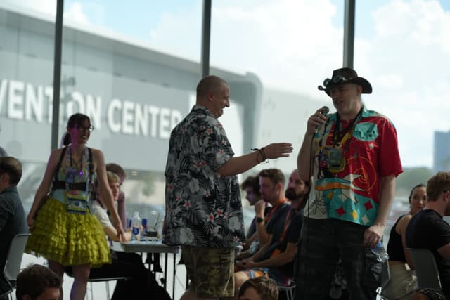 A group of people gathered indoors, with two individuals standing and conversing in the foreground. One person is holding a microphone, and others are seated at tables in the background. The setting appears to be a convention center, as indicated by the large windows and signage