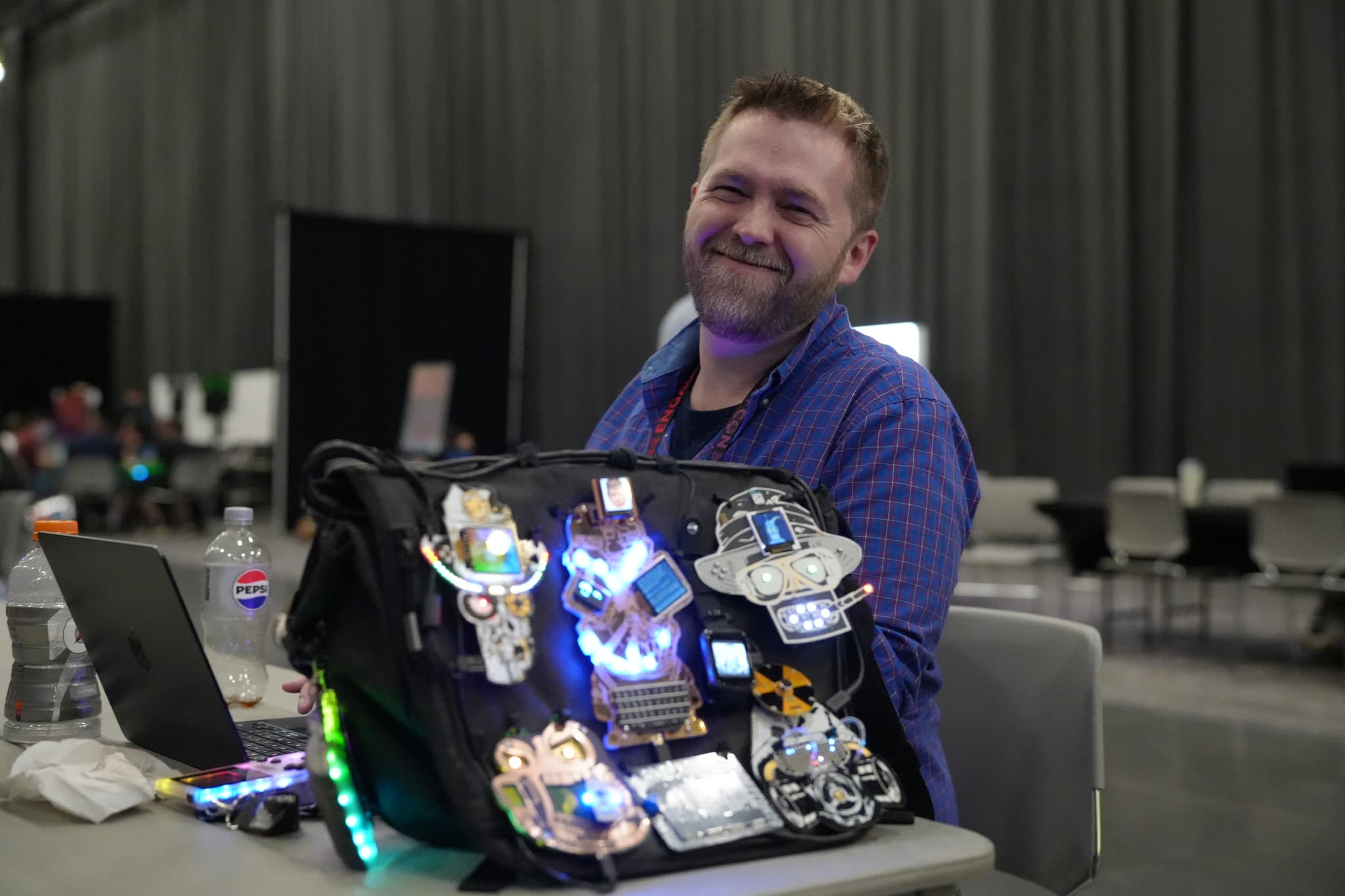 A man sitting at a table with a laptop, displaying a collection of illuminated badges on a bag