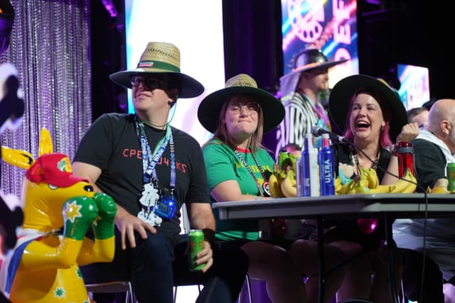 A group of people wearing hats and lanyards sit at a table with colorful decorations and drinks, while a person in a kangaroo costume stands nearby