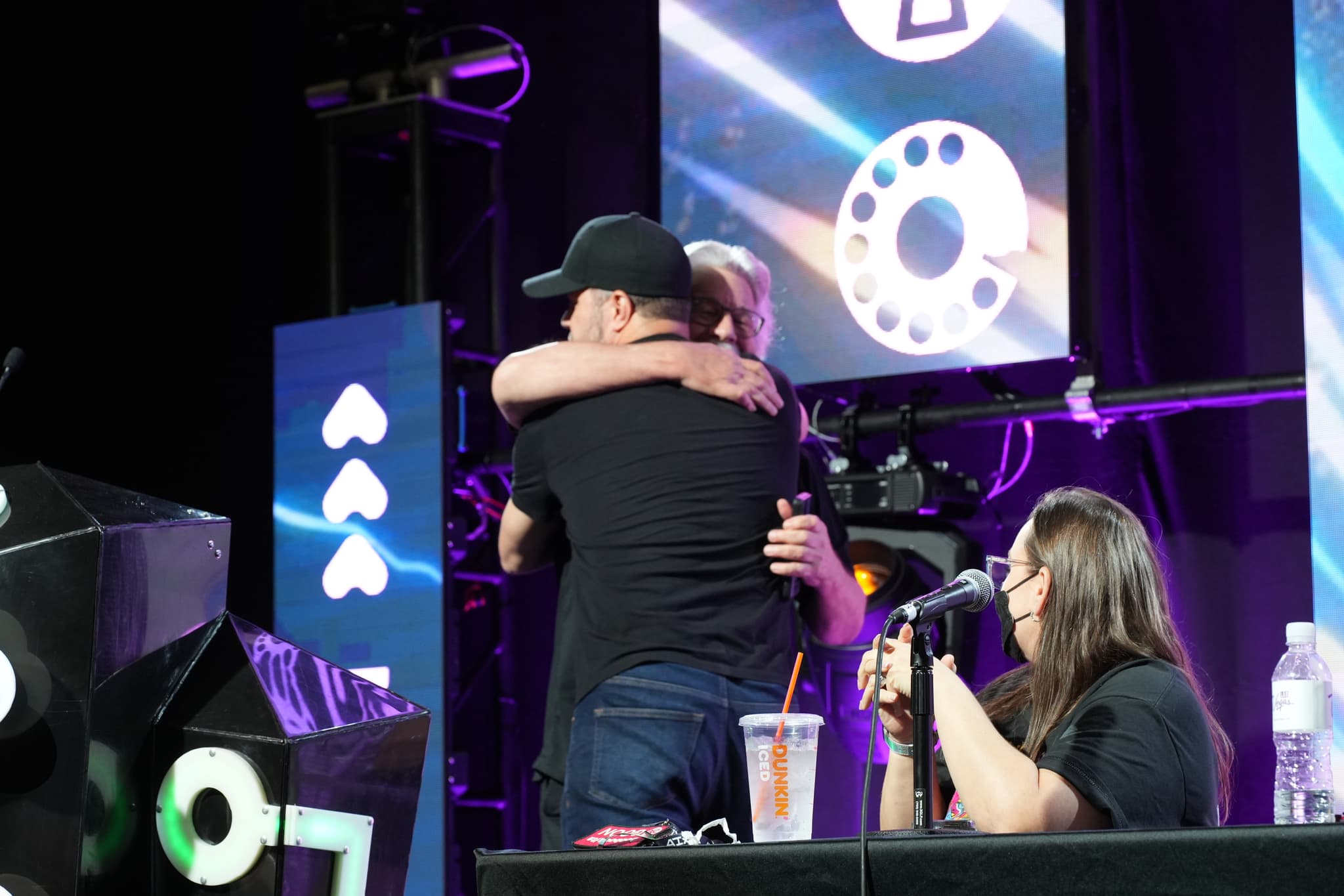 Two people embracing on a stage with a woman sitting at a table nearby, holding a microphone. The background features large screens with various symbols and lights