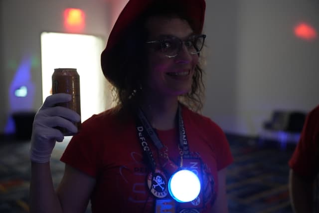 A person wearing a red hat and glasses, holding a can in one hand, with a glowing blue light on their chest. The background is dimly lit with some colorful lights