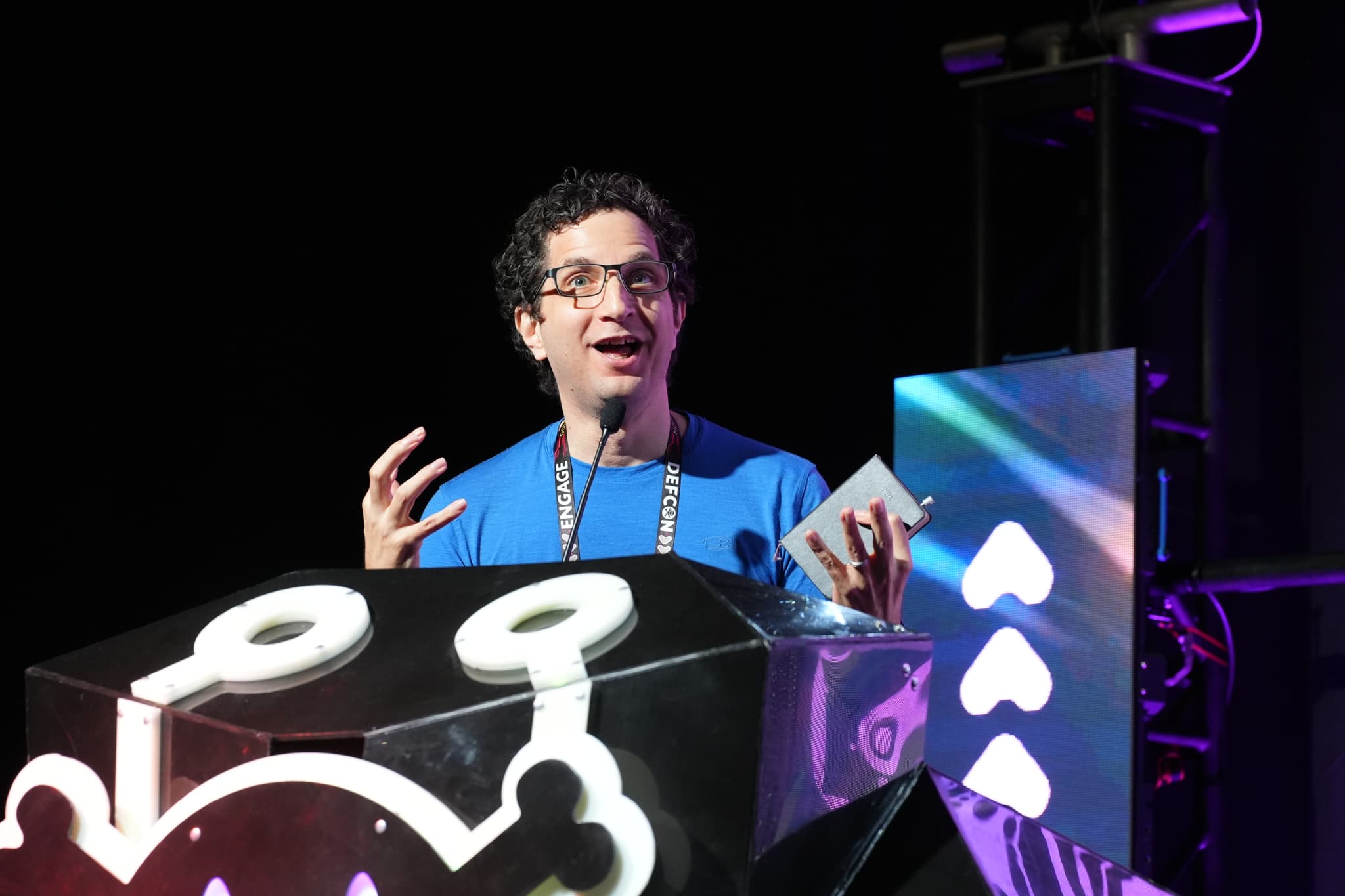 A person wearing glasses and a blue shirt is speaking animatedly at a podium decorated with a cartoonish design. The background features colorful lighting and geometric shapes