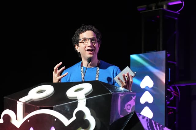 A person wearing glasses and a blue shirt is speaking animatedly at a podium decorated with a cartoonish design. The background features colorful lighting and geometric shapes