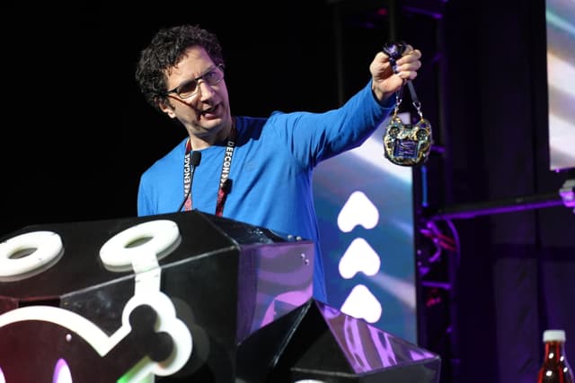 A person wearing glasses and a blue shirt is holding up a device while standing behind a podium with a stylized design. The background features stage lighting and a screen with geometric shapes