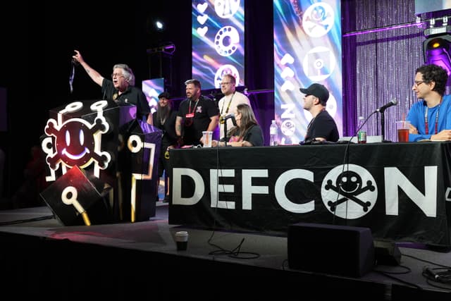 A group of people on a stage at DEFCON, with one person standing and speaking at a podium decorated with a hacker-themed logo