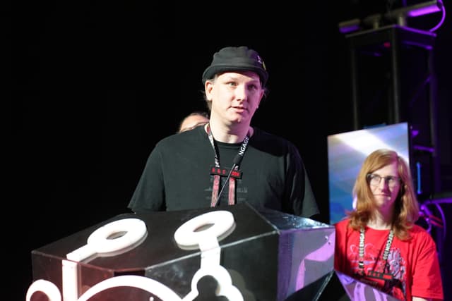 A man wearing a black hat and a black shirt stands next to a woman with glasses and red hair. They are in front of a large black and white object with a design on it, and the background is dark with some lighting equipment visible