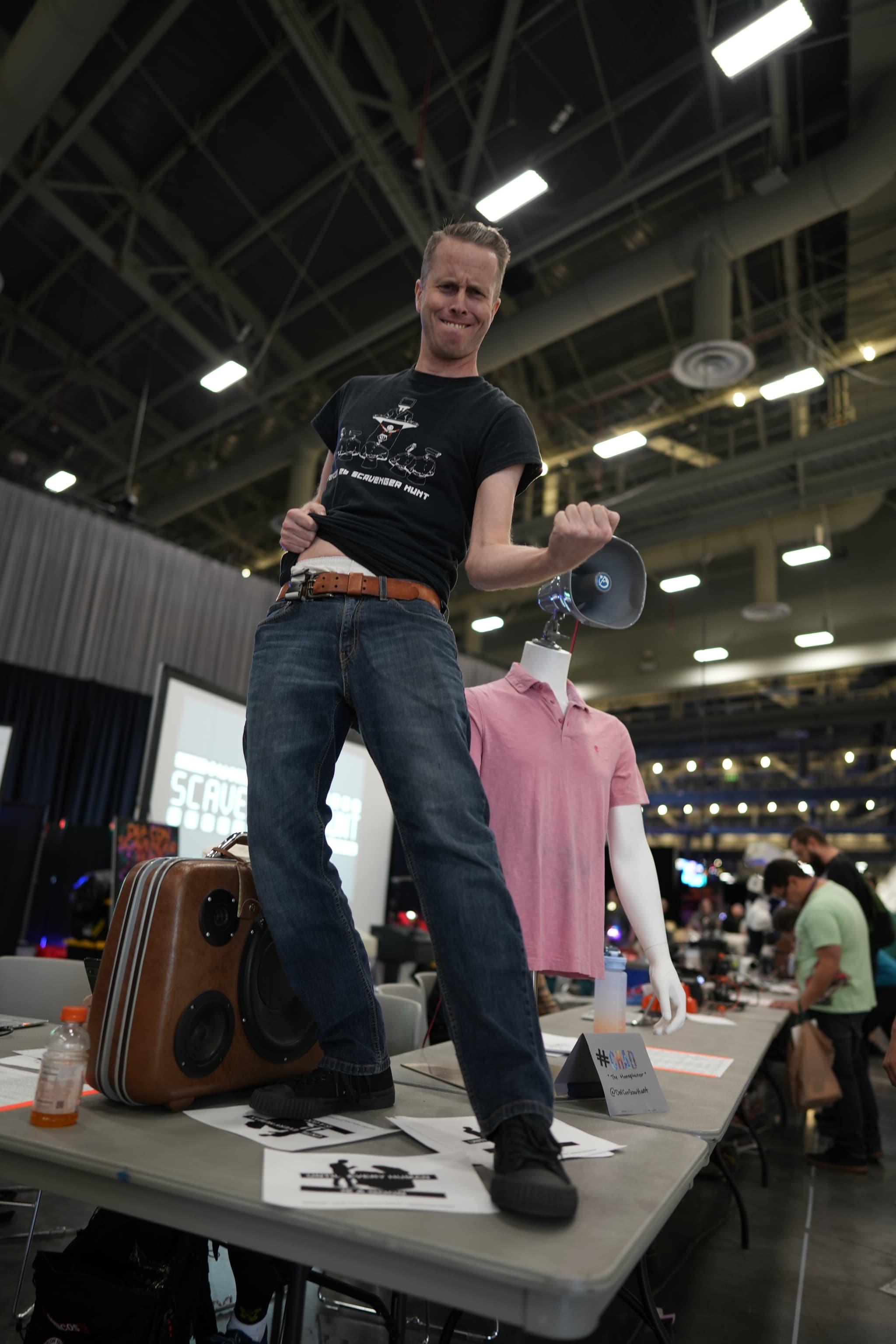 A person standing on a table, holding a megaphone and posing energetically, with various items and a mannequin in the background. The setting appears to be an indoor event or convention