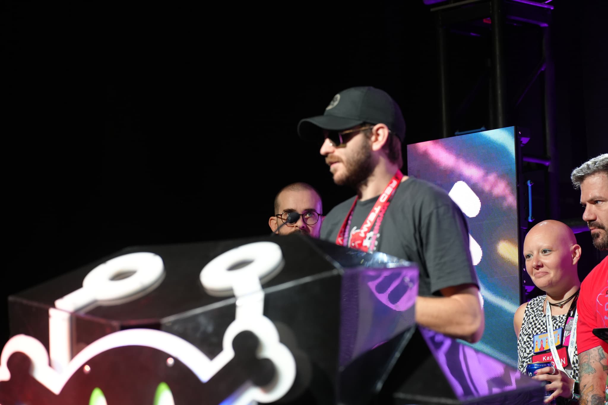 A group of people standing behind a podium with a cartoonish design, one person wearing a cap and sunglasses, and colorful lights in the background