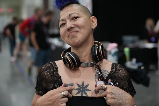 A person with a mohawk hairstyle and headphones around their neck is pointing to a star-shaped tattoo on their chest. The individual is wearing a black lace top and has additional tattoos on their arms. The background appears to be an indoor event with other people and booths