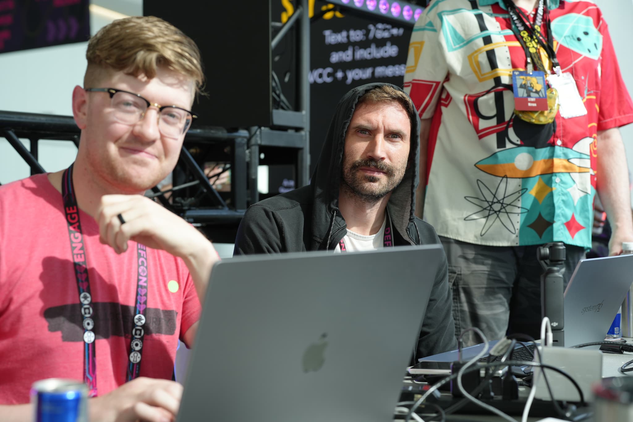 Two men sitting at a table with laptops, one wearing a red shirt and glasses, the other in a black hoodie. A third person, partially visible, stands nearby wearing a colorful shirt