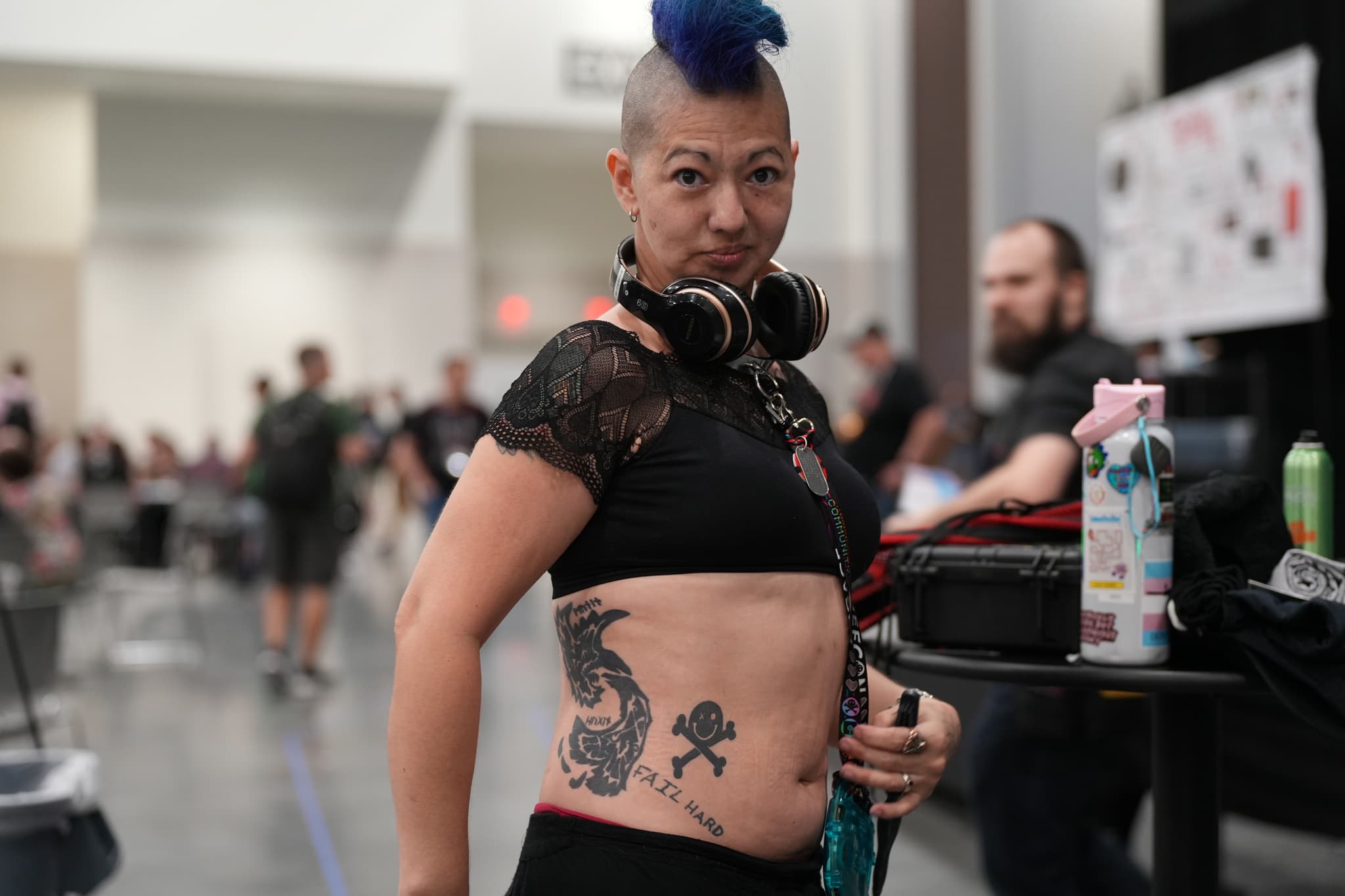 A person with a blue mohawk, wearing headphones around their neck, displays tattoos on their torso in an indoor setting with other people in the background