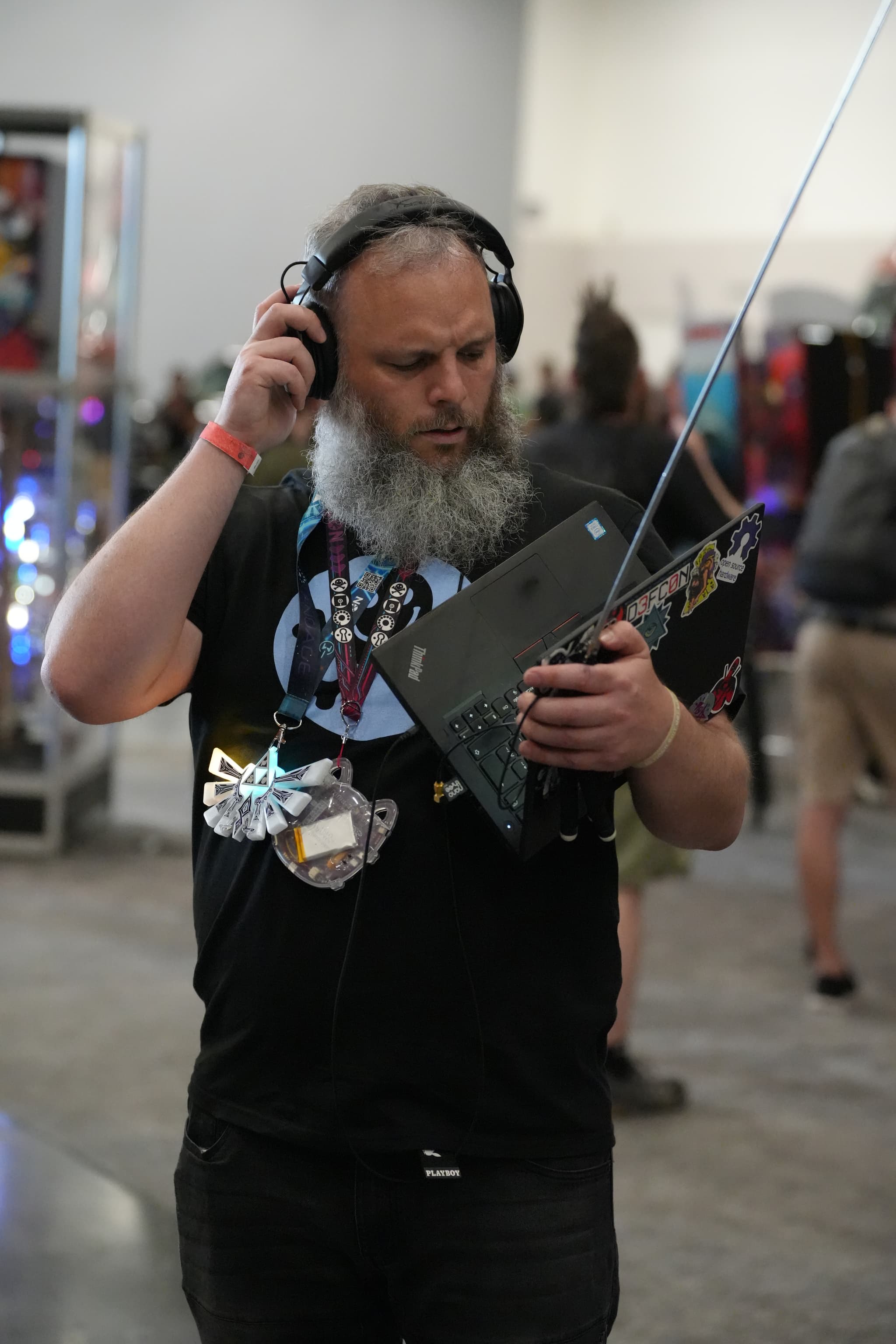 A man with a beard wearing headphones and a black t-shirt is holding a laptop with an antenna attached to it. He appears to be in a busy indoor setting with other people and various equipment in the background