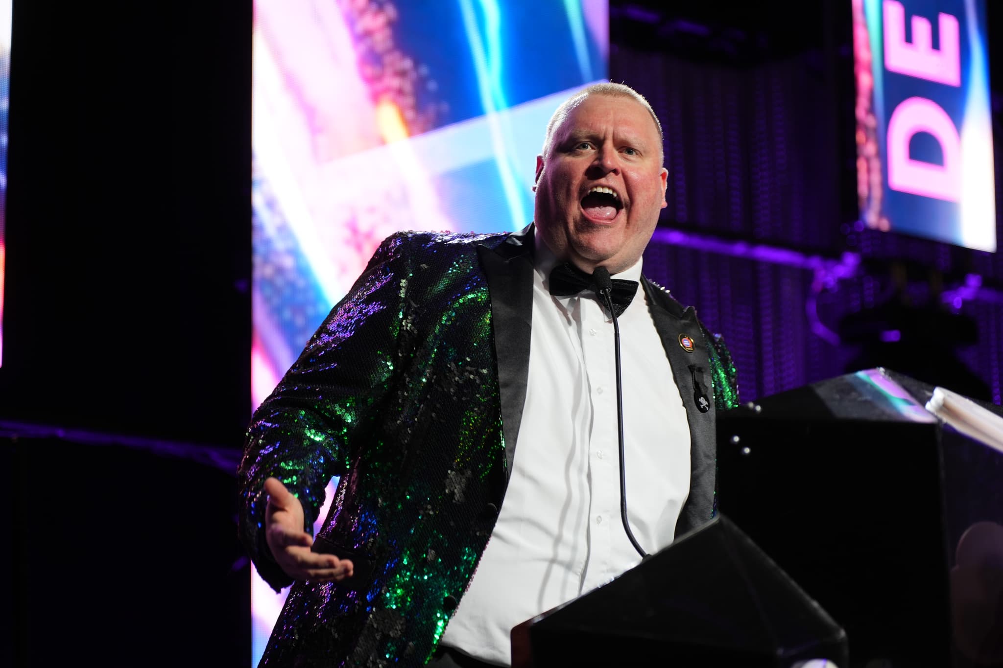 A man in a sparkly green jacket and bow tie is speaking passionately at a podium with colorful lights in the background