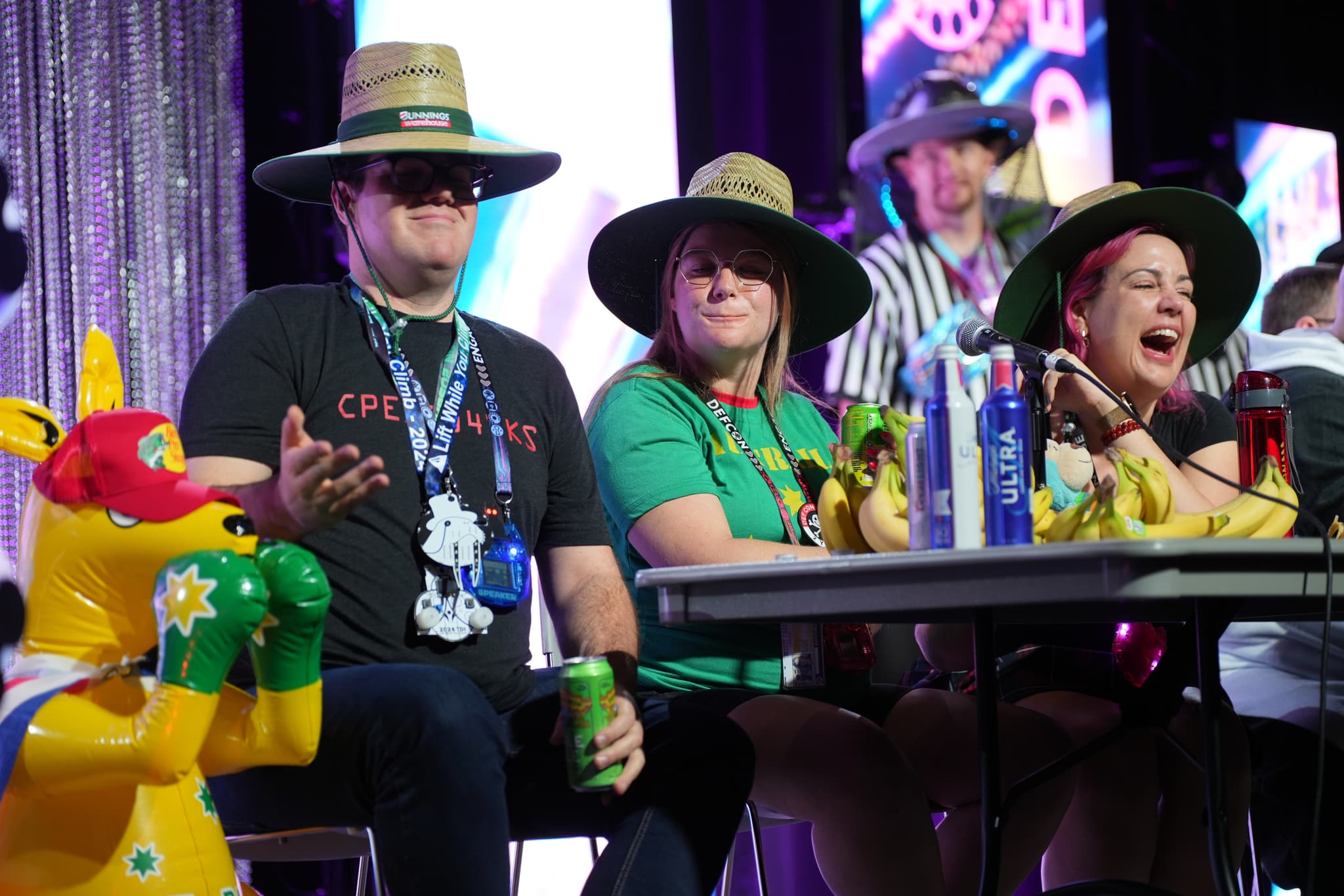 A group of people wearing wide-brimmed hats are sitting at a table with various items, including bananas and drinks. One person is holding a can, and there is a colorful inflatable kangaroo nearby. The background features a stage with lights and decorations