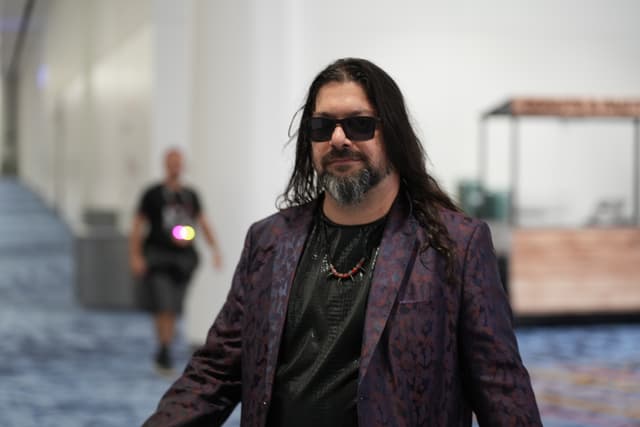 A man with long hair and sunglasses wearing a dark purple jacket and black shirt, standing in an indoor setting with a blurred background