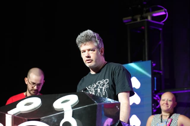 A man with gray hair stands behind a podium with a microphone, flanked by two other individuals, one on each side. The background features stage lighting and equipment