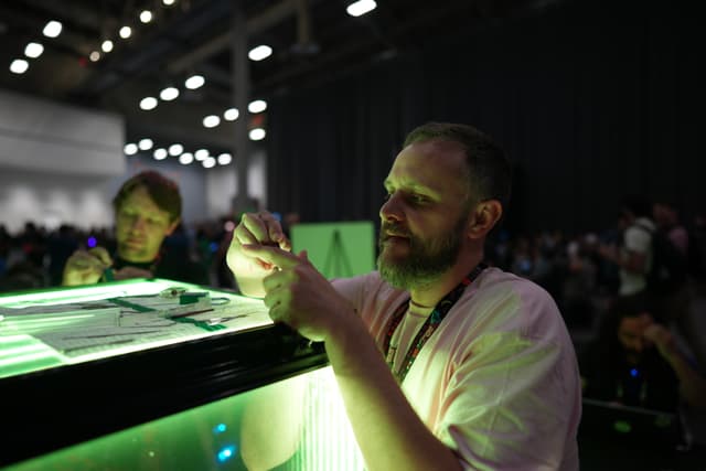 A man is working on a small object under green lighting, with another person in the background in a large, dimly lit room