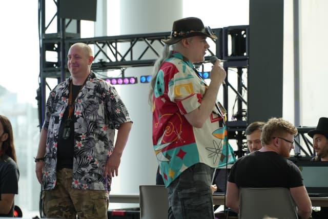 Two men in colorful shirts, one holding a microphone, stand in front of a group of seated people in a well-lit room with a metal framework and lighting equipment in the background