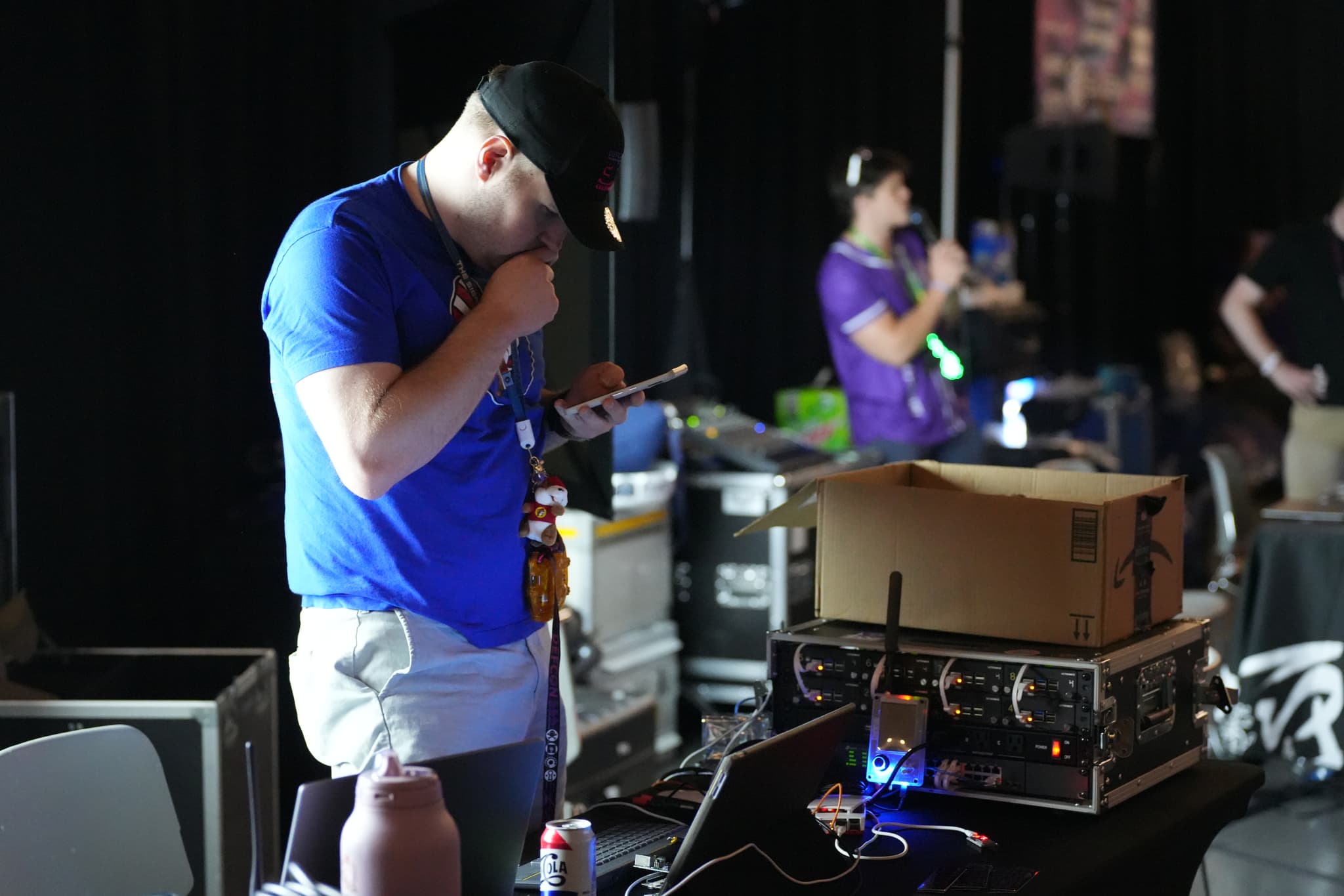 A person in a blue shirt and cap is standing at a table with electronic equipment, looking at a tablet. Another person in the background is speaking into a microphone