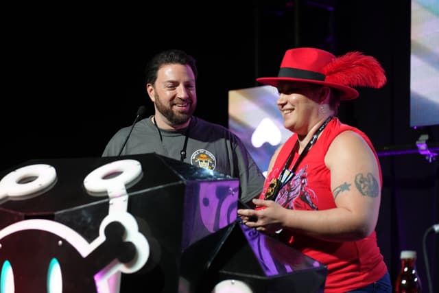 Two people standing behind a podium, one wearing a red hat with feathers and a red shirt, and the other wearing a gray shirt. Both are smiling and appear to be engaged in conversation